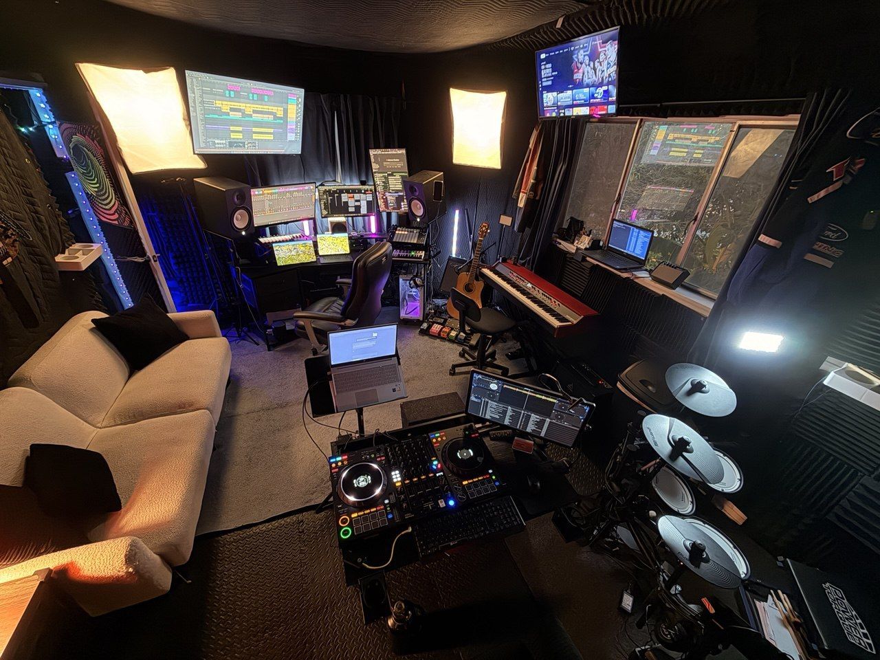 Elevated wide shot, full studio with DAW, DJ deck, drums, couch, multiple screens